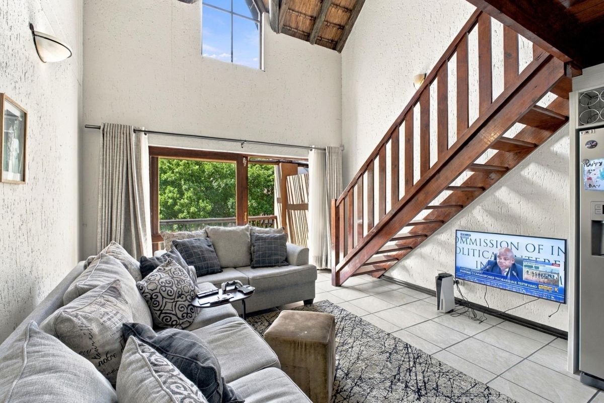 Spacious double-volume living area featuring exposed timber rafters, a wooden staircase, tiled flooring, and large windows providing natural light and garden access.
