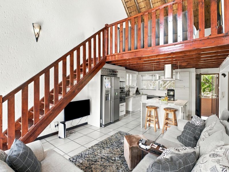 Open-plan living area featuring a kitchen with an island, tiled floors, and a wooden staircase leading to a mezzanine level with a thatched roof.