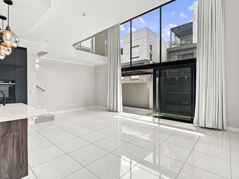 Spacious open-plan living area featuring double-volume windows, sleek porcelain tiles, and a modern kitchen with integrated appliances, flowing seamlessly to the outdoor patio.
