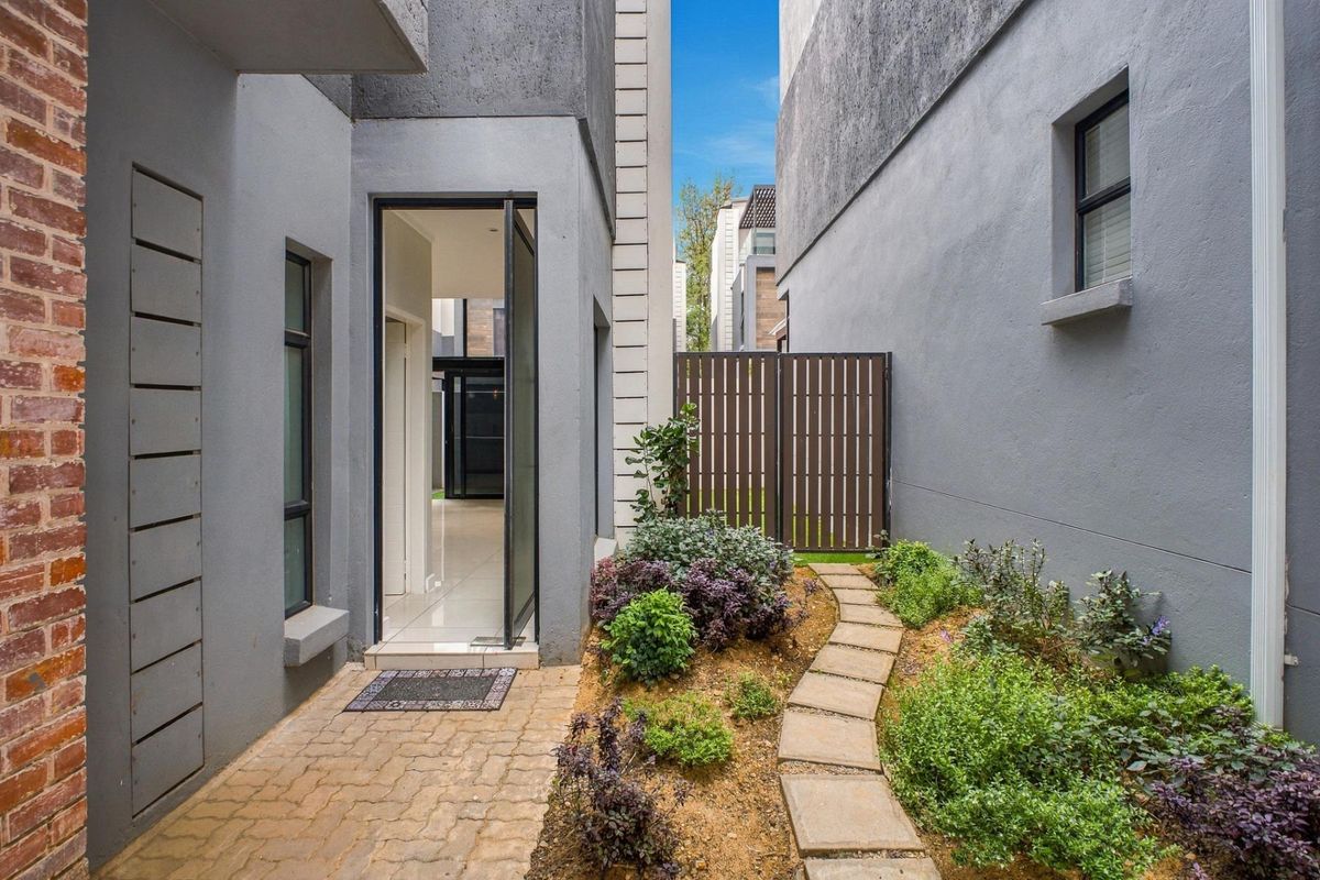 A well-maintained rear exterior area featuring a paved walkway, landscaped garden beds, and a private entrance leading into the townhouse.