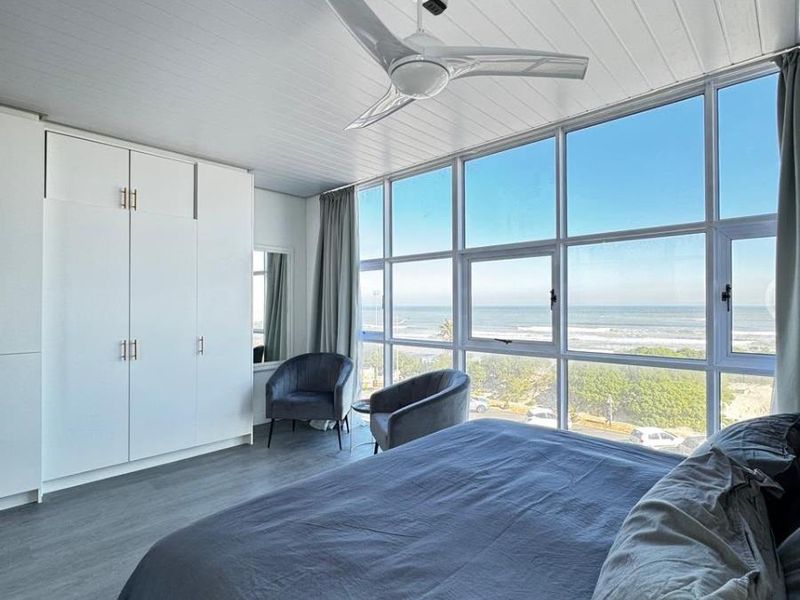 A bright bedroom featuring built-in cupboards, a modern ceiling fan, and expansive windows offering panoramic ocean views.