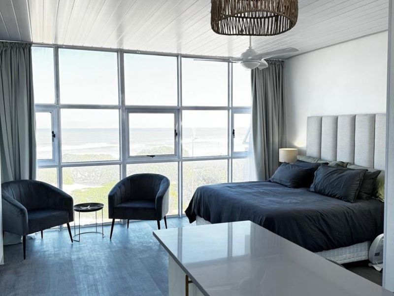 Spacious bedroom featuring large floor-to-ceiling windows with ocean views, a modern kitchen island, and a contemporary tongue and groove ceiling.