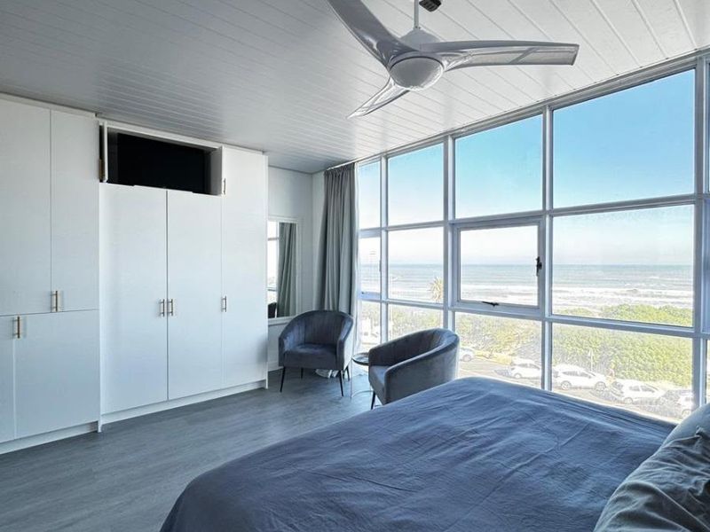 A bright bedroom featuring built-in cupboards, a modern ceiling fan, and expansive floor-to-ceiling windows offering panoramic ocean views.
