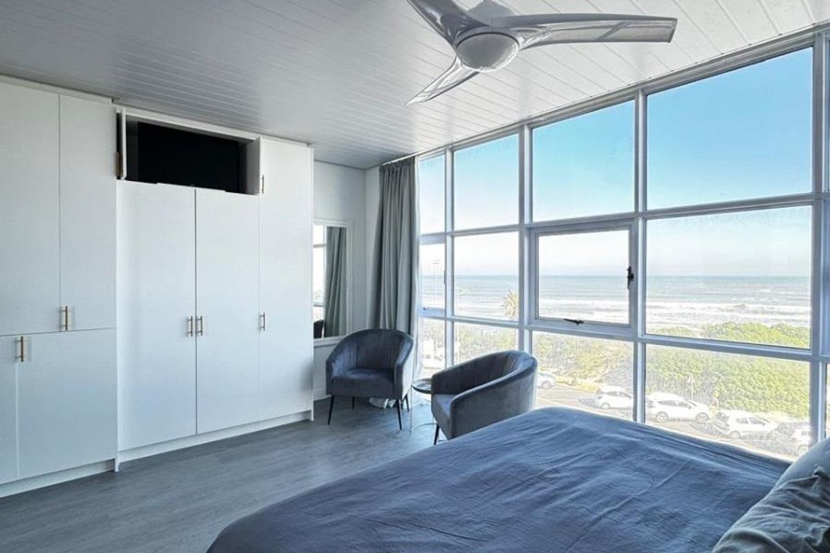 A bright bedroom featuring built-in cupboards, a modern ceiling fan, and expansive floor-to-ceiling windows offering panoramic ocean views.