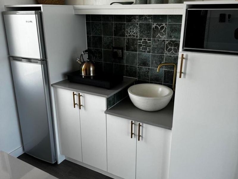 Compact kitchen area featuring white cabinetry, stone countertops, a modern vessel sink with brass faucet, and a decorative patterned tile backsplash.