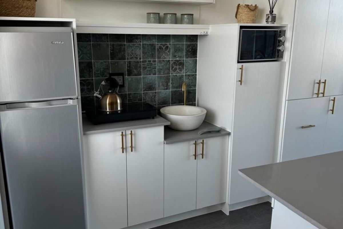 Modern compact kitchen featuring white cabinetry, grey stone countertops, a decorative tiled backsplash, and a stylish vessel sink with brass fixtures.