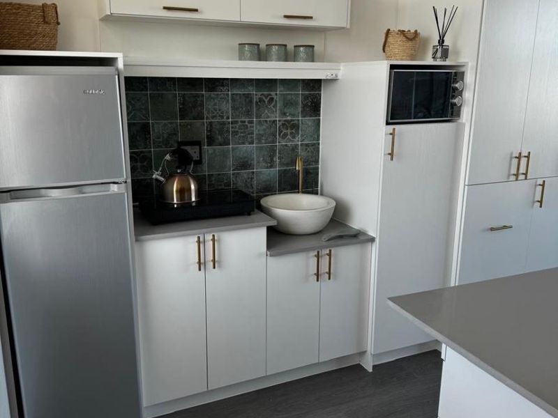 Modern compact kitchen featuring white cabinetry, grey stone countertops, a decorative tiled backsplash, and a stylish vessel sink with brass fixtures.