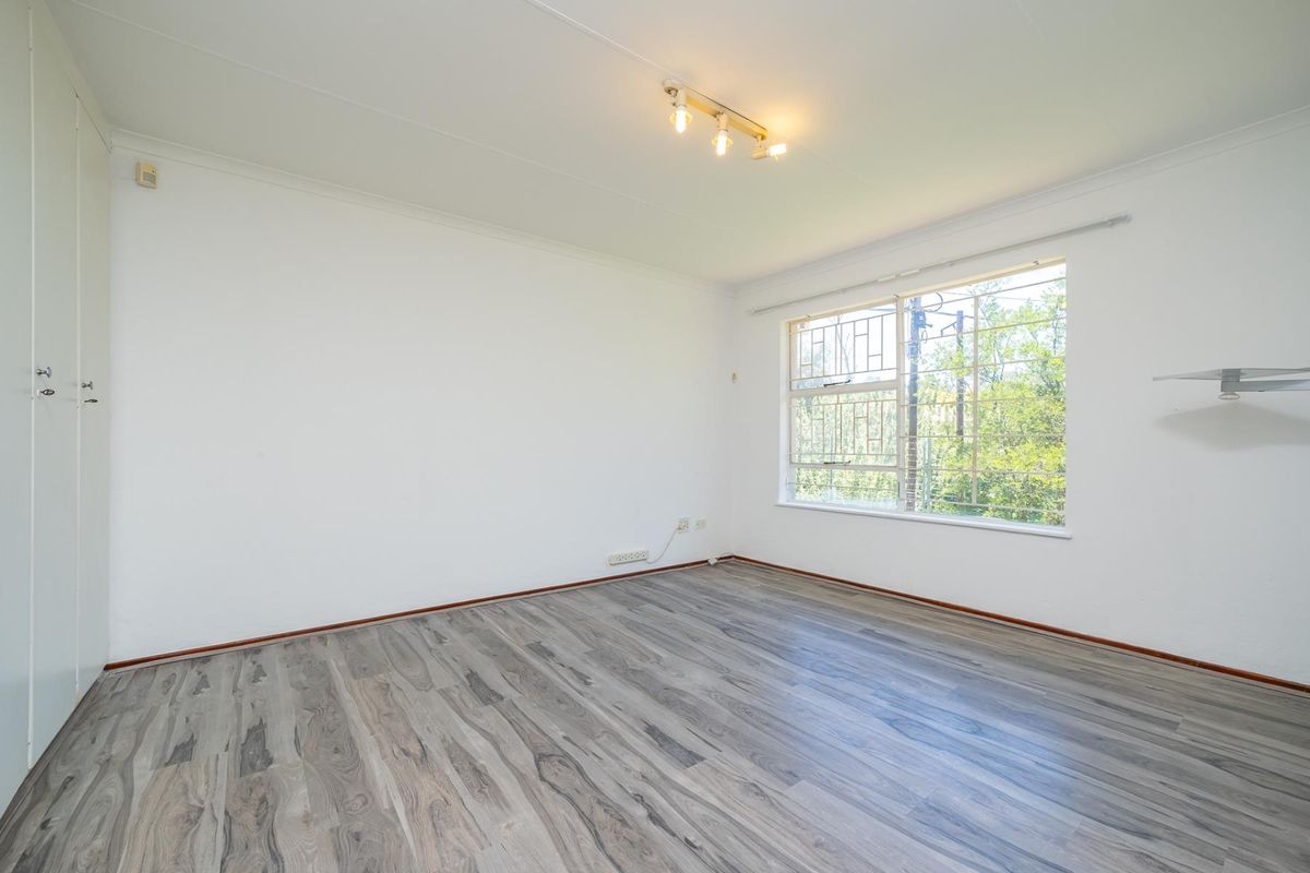 A bright, empty bedroom featuring modern laminate flooring, built-in cupboards, and a window with security bars, offering a clean and neutral living space.