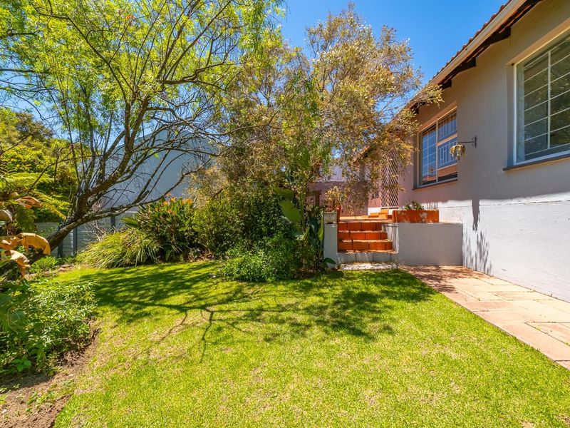 A sunny rear garden featuring a well-maintained lawn, mature trees, and a paved walkway leading to tiled steps against the townhouse exterior.