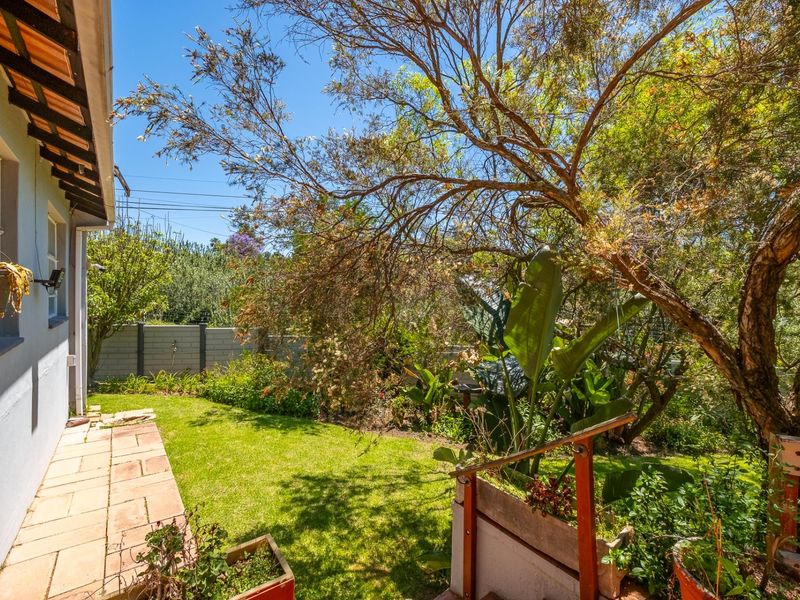 A rear garden area featuring a paved patio, a well-maintained lawn, and mature trees, bordered by a perimeter wall.