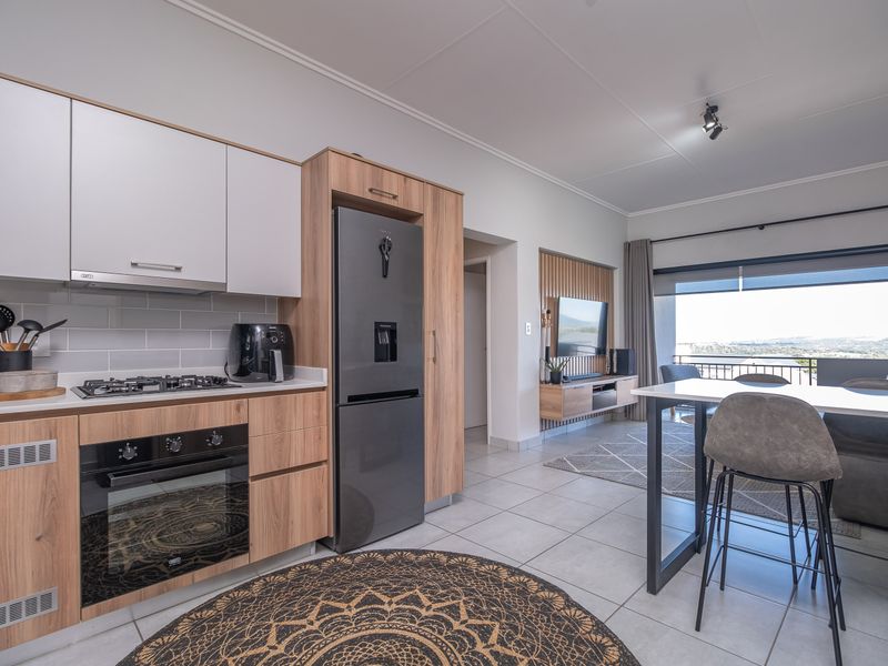 Modern open-plan living area featuring a contemporary kitchen with wood-finish cabinetry, gas hob, and integrated oven, flowing seamlessly to a balcony with scenic views.