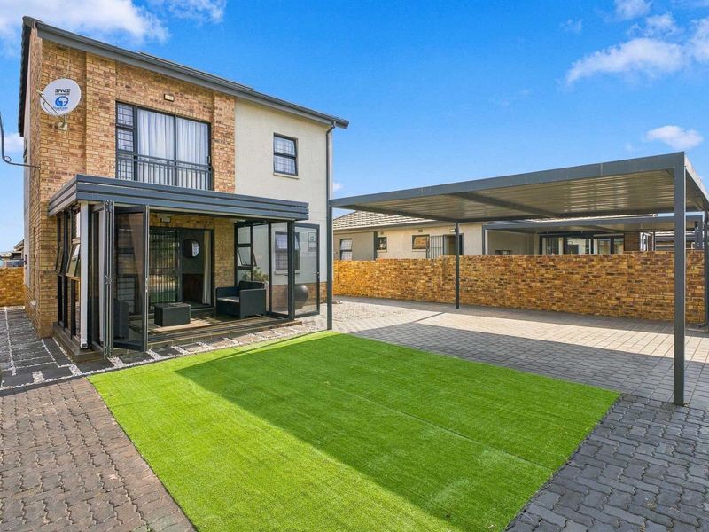 Rear exterior view featuring a paved driveway, carport, artificial lawn, and an enclosed patio with folding glass doors attached to the brick townhouse.