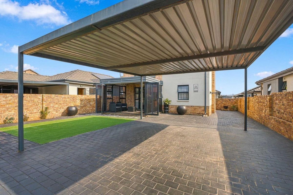A well-maintained rear exterior featuring a large metal carport, paved driveway, artificial lawn, and an enclosed patio area with brick boundary walls.