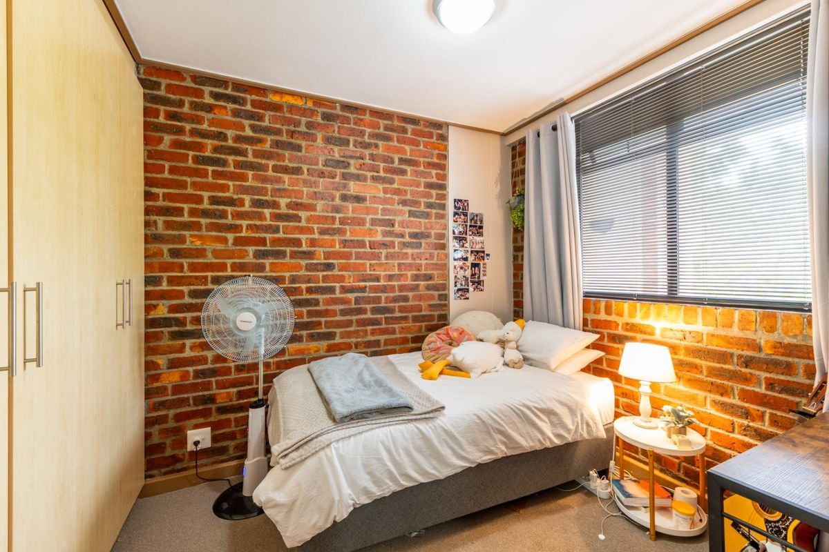 A cozy bedroom featuring an exposed brick feature wall, built-in wardrobe storage, and large windows with venetian blinds.