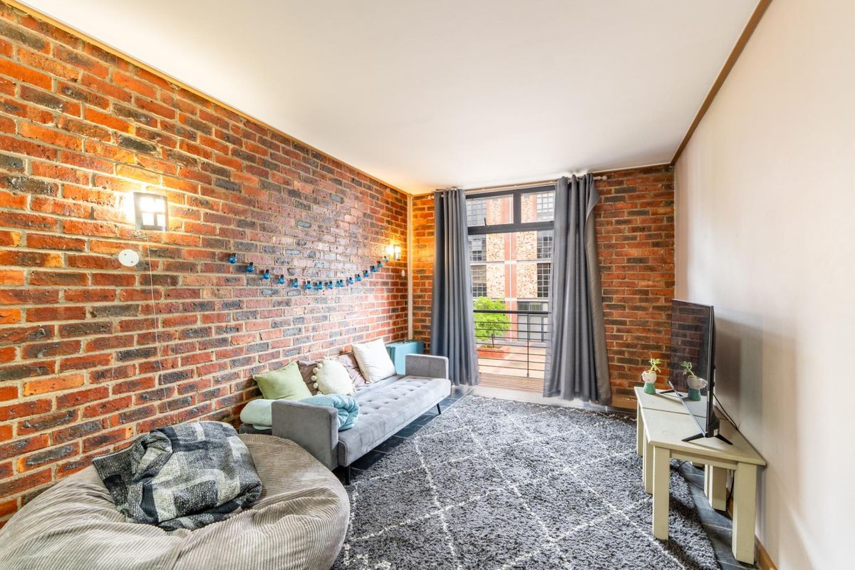A cozy living room featuring an exposed brick wall, large window, and direct access to a balcony, offering a warm and industrial aesthetic.