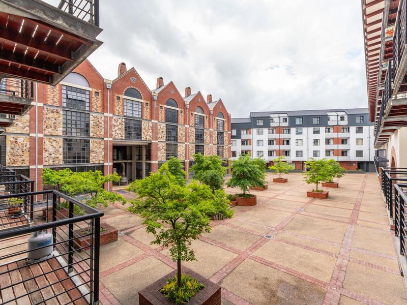 A well-maintained central courtyard area featuring brick paving, raised planters with trees, and surrounding multi-story apartment building facades with industrial-style architecture.