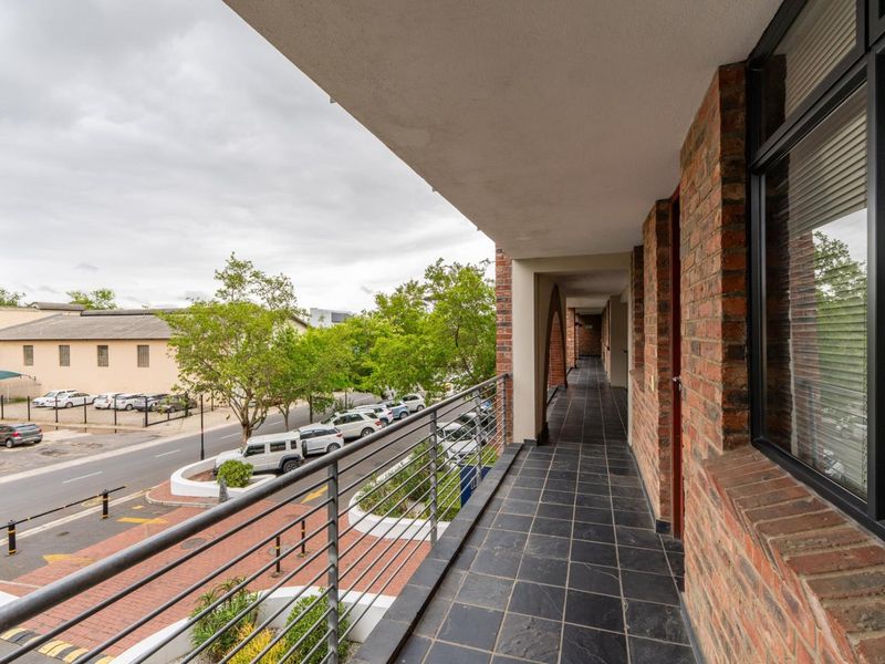 A covered balcony featuring exposed brick walls, dark tiled flooring, and a metal safety railing, overlooking the street and surrounding neighborhood.