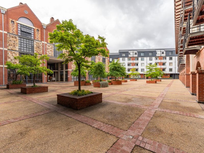 A spacious, well-maintained paved courtyard featuring brick planters and mature trees, surrounded by multi-story apartment buildings with exposed brick and modern architectural details.
