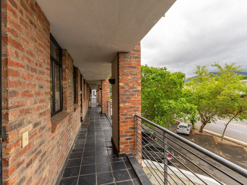 A covered balcony featuring exposed brick walls, dark tiled flooring, and a metal safety railing overlooking the street and surrounding greenery.