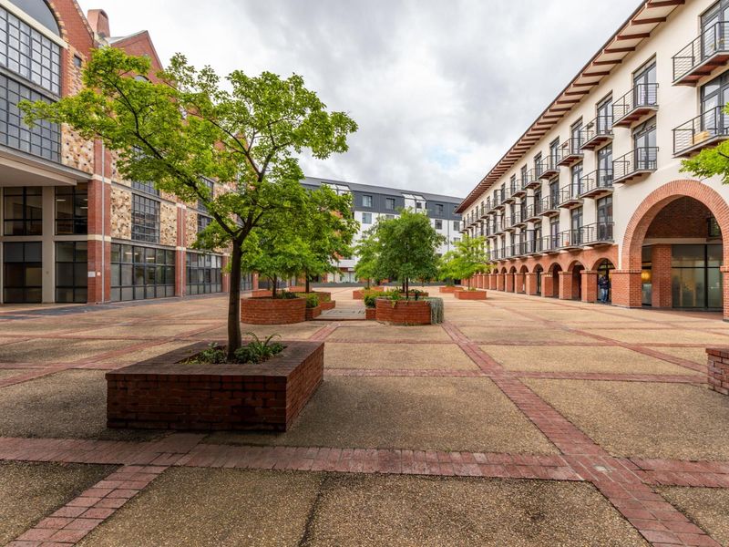 A spacious, well-maintained paved courtyard area between apartment buildings, featuring brick planters, mature trees, and architectural arched entryways.