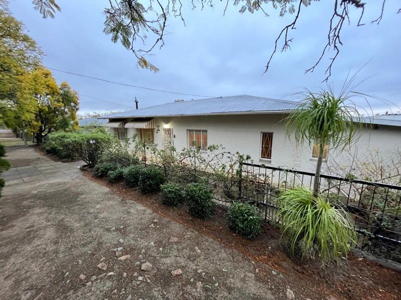 Front elevation of a single-story house featuring a corrugated metal roof, security gates, and established garden beds along a concrete driveway.