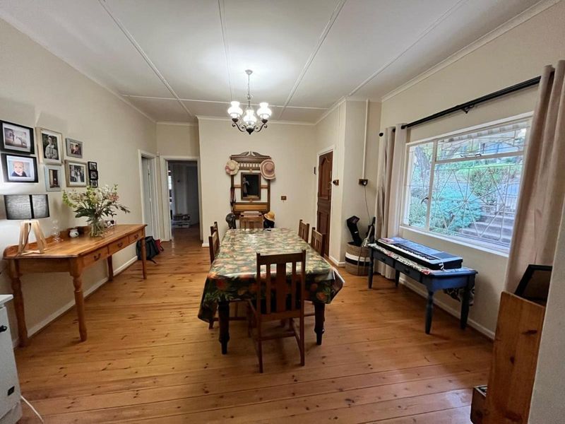A spacious dining room featuring warm timber flooring, a classic ceiling light fixture, and a large window with security bars overlooking the garden.