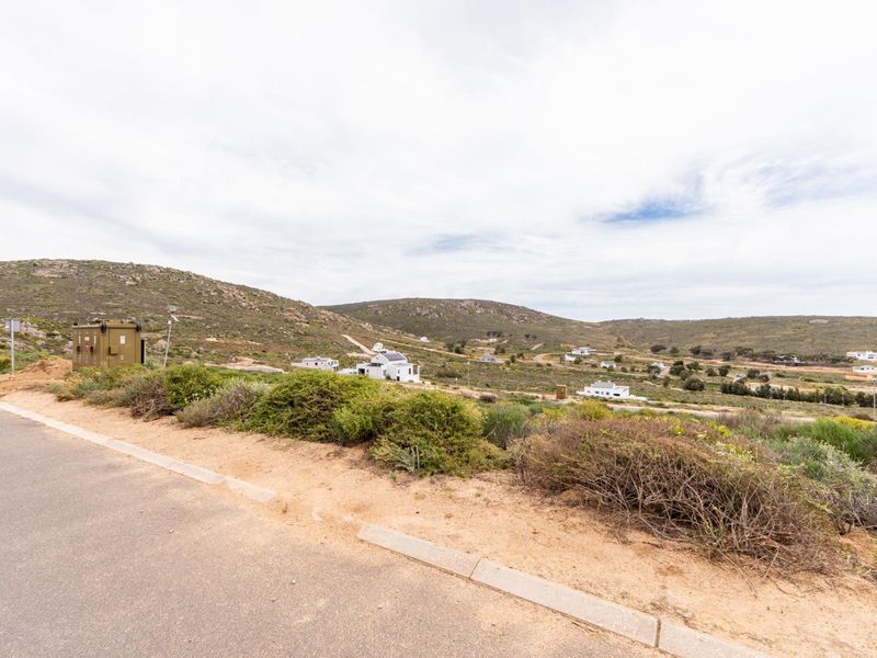 A street-level view of a residential development area featuring paved road access, concrete curbing, and surrounding natural landscape with distant homes.
