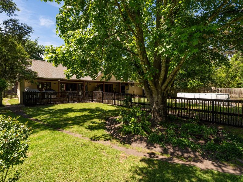 A spacious rear garden featuring a well-maintained lawn, mature trees, and a fenced-off area with a covered patio attached to the main house.