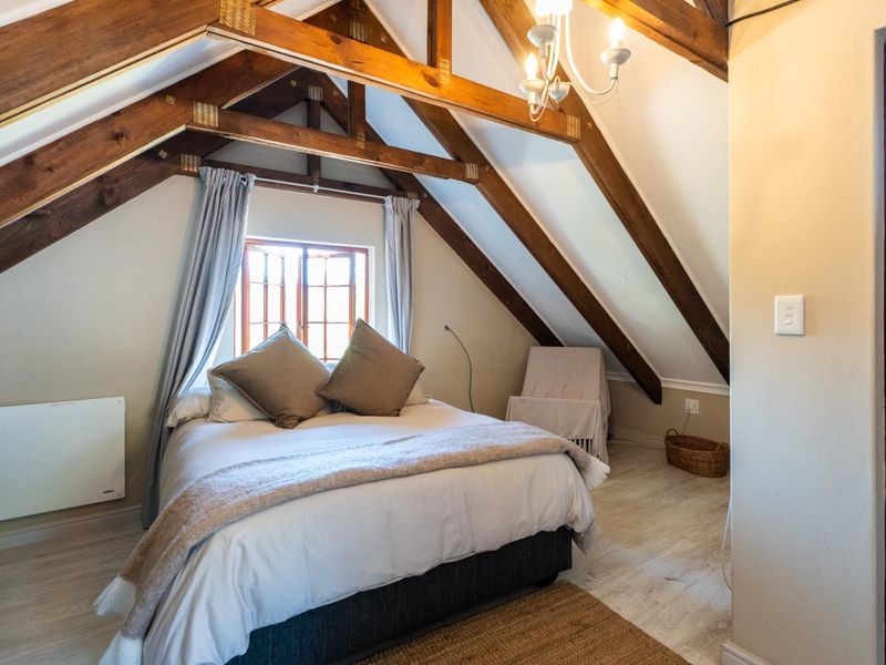 Cozy attic bedroom featuring exposed timber roof trusses, vaulted ceilings, and a large timber-framed window, creating a warm and inviting atmosphere.