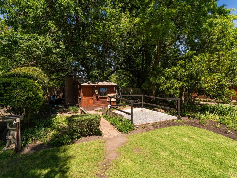 A lush garden area featuring a wooden shed, a gravel patio space with metal railings, and a brick pathway surrounded by mature trees.
