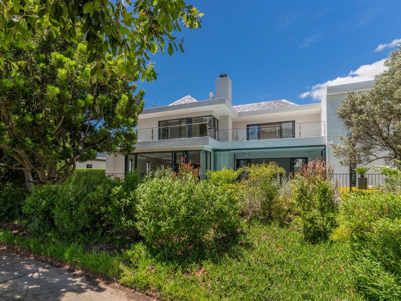 Rear view of a modern two-story house featuring a balcony with glass balustrades, large sliding glass doors, and a well-maintained garden.