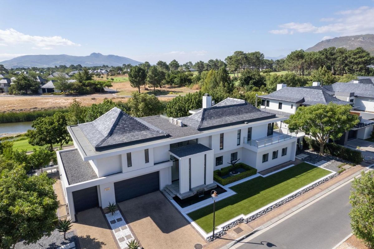 Aerial view of a modern luxury residence featuring a slate roof, double garage, manicured lawn, and balcony, situated in a scenic estate setting.