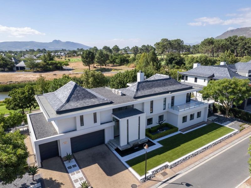 Aerial view of a modern luxury residence featuring a slate roof, double garage, manicured lawn, and balcony, situated in a scenic estate setting.