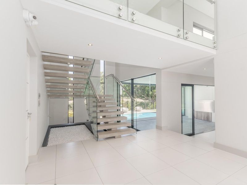 Spacious modern entrance hall featuring a striking floating staircase with glass balustrades, large format floor tiles, and seamless views toward the outdoor pool area.