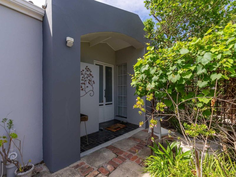 A charming covered entrance featuring a dark grey arched facade, tiled porch, and a white framed glass door, surrounded by lush garden greenery.