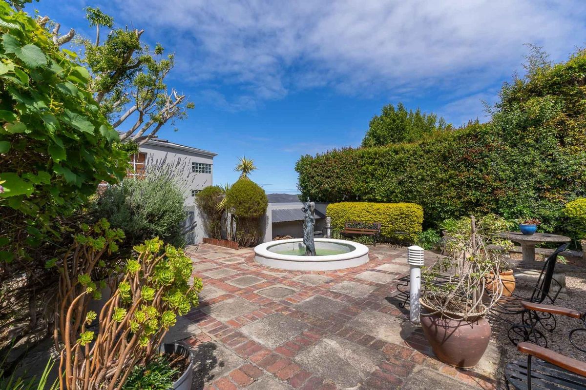 A private rear garden featuring a central stone fountain, brick-paved patio area, and established perimeter hedging.