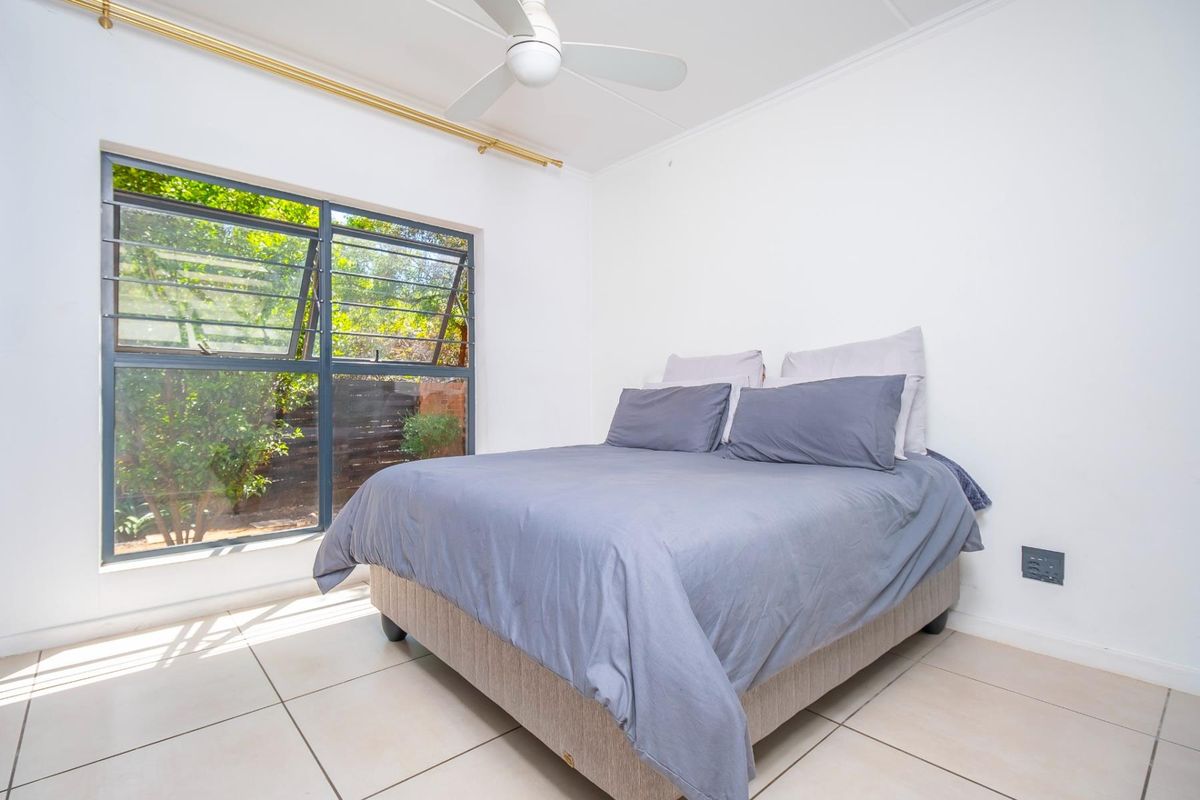 A bright bedroom featuring tiled flooring, a ceiling fan, and large windows with security bars overlooking the garden.