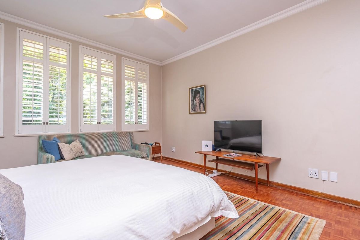 A well-lit bedroom featuring classic parquet flooring, elegant plantation shutters, and a ceiling fan, offering a comfortable and inviting living space.