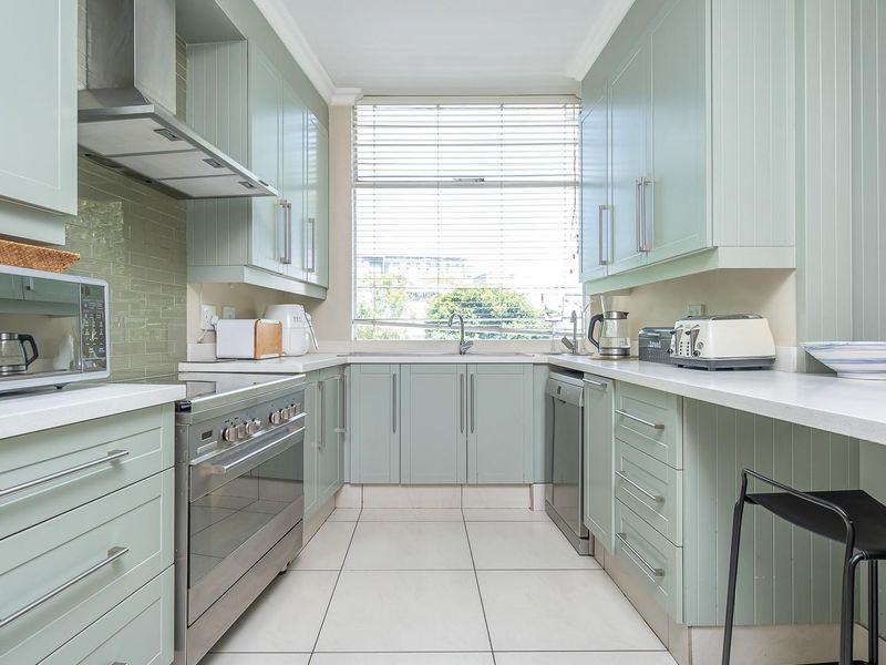 A well-maintained kitchen featuring light green cabinetry, stone countertops, stainless steel appliances, and a tiled backsplash with ample natural light.