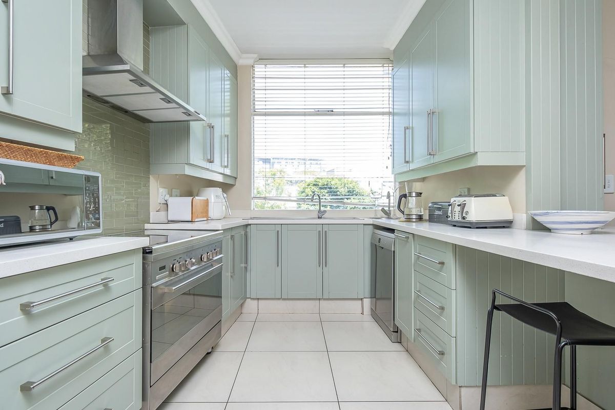 A well-maintained kitchen featuring light green cabinetry, stone countertops, stainless steel appliances, and a tiled backsplash with ample natural light.