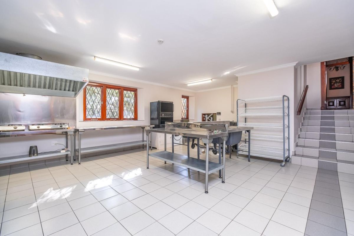 A functional commercial-style kitchen featuring stainless steel work surfaces, a built-in extractor hood, tiled flooring, and direct access to a staircase.