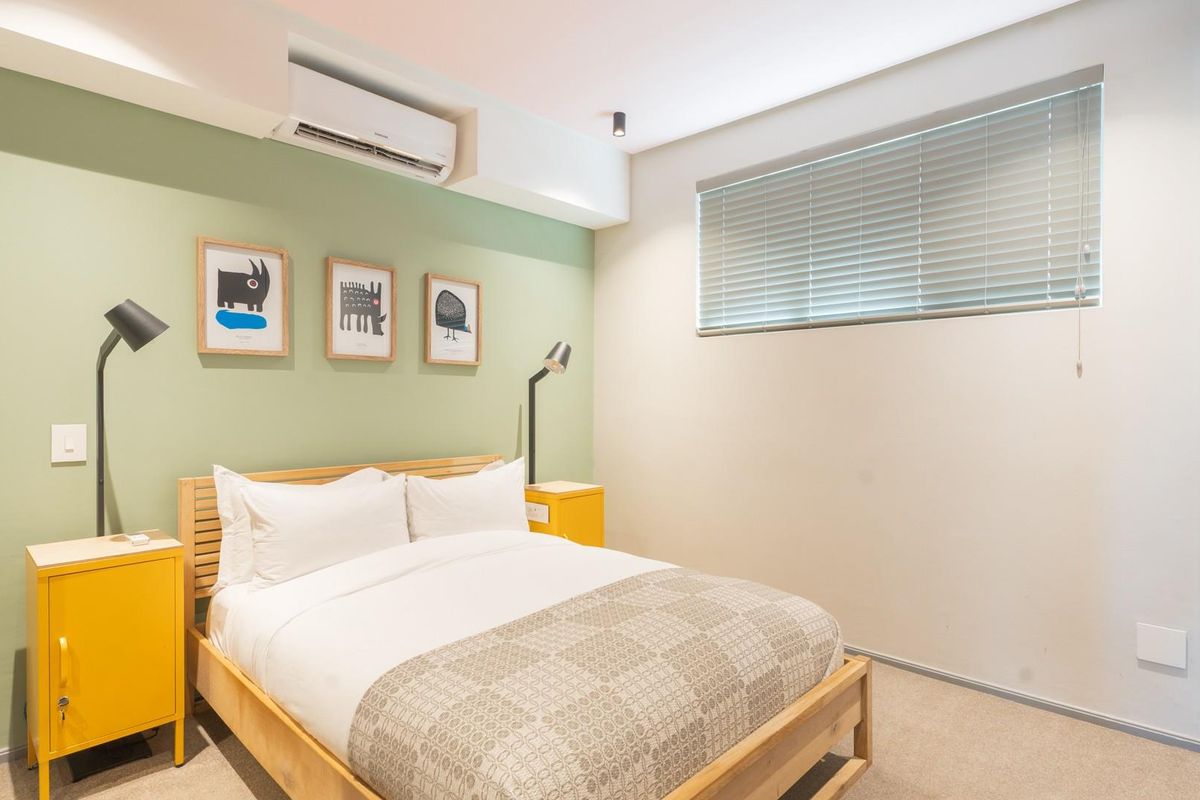A well-maintained bedroom featuring a soft green accent wall, integrated air conditioning unit, and horizontal window blinds.