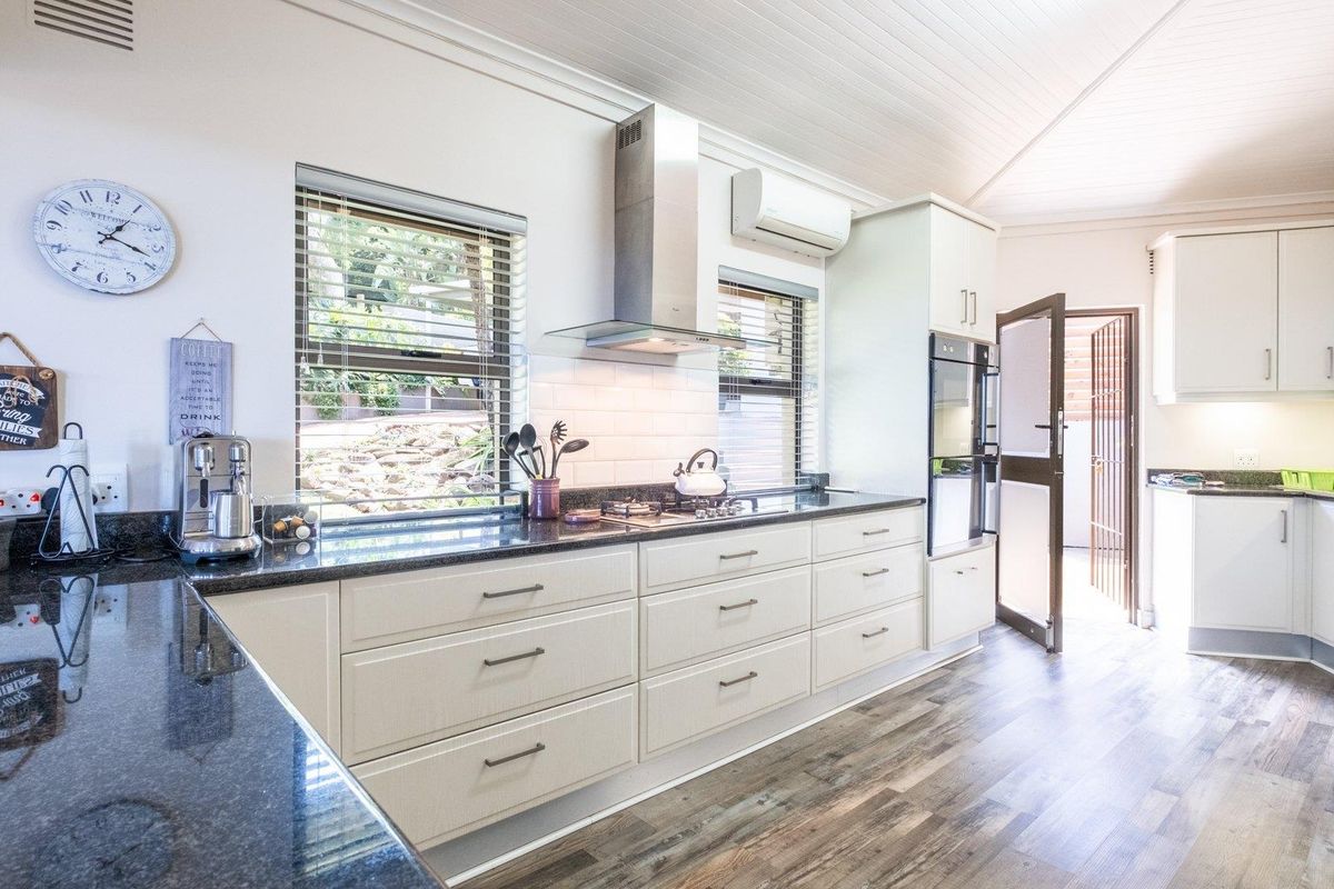 A modern, well-lit kitchen featuring white cabinetry, dark granite countertops, a stainless steel extractor fan, built-in oven, and wood-look flooring.
