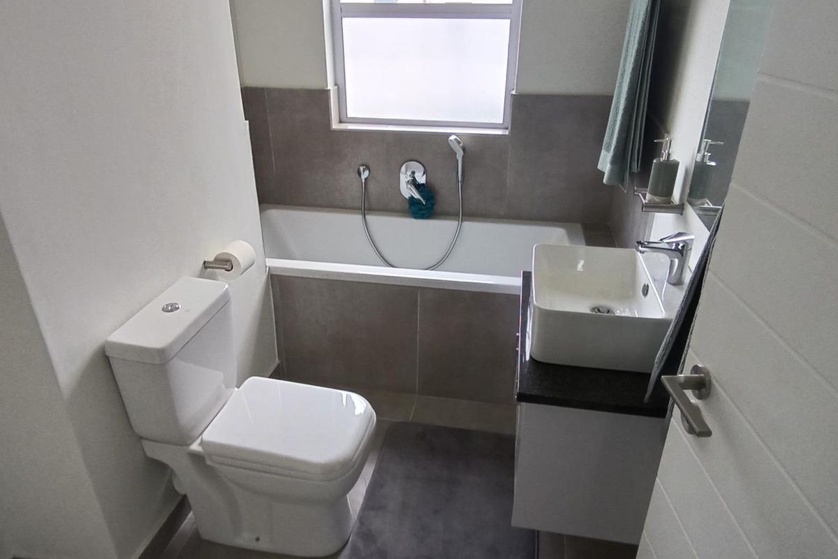 A modern bathroom featuring a built-in bathtub, a vessel sink vanity, and a toilet, finished with neutral wall tiles and a frosted window.