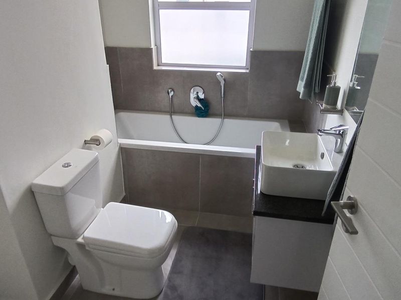 A modern bathroom featuring a built-in bathtub, a vessel sink vanity, and a toilet, finished with neutral wall tiles and a frosted window.