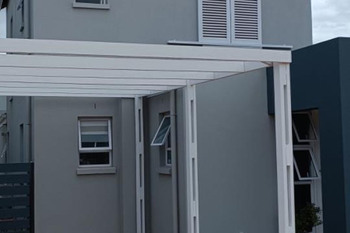 A modern two-story residential facade featuring a paved driveway, a white pergola structure, and grey exterior wall finishes with casement windows.
