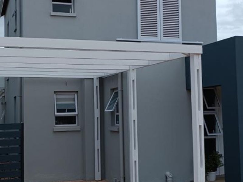 A modern two-story residential facade featuring a paved driveway, a white pergola structure, and grey exterior wall finishes with casement windows.