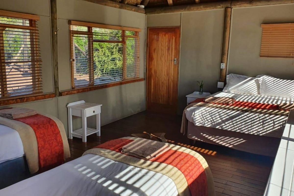 A rustic bedroom featuring timber flooring, exposed structural timber poles, and large windows with venetian blinds, offering a warm and natural aesthetic.