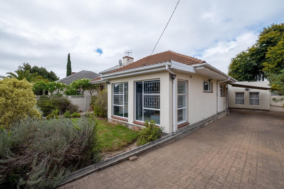 Rear view of the property featuring a paved driveway, landscaped garden area, and a tiled roof structure with security bars on the windows.