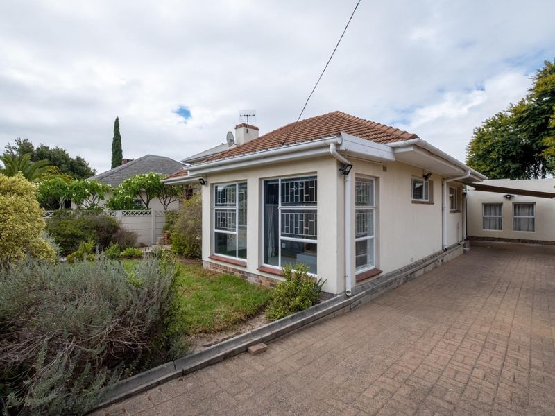 Rear view of the property featuring a paved driveway, landscaped garden area, and a tiled roof structure with security bars on the windows.